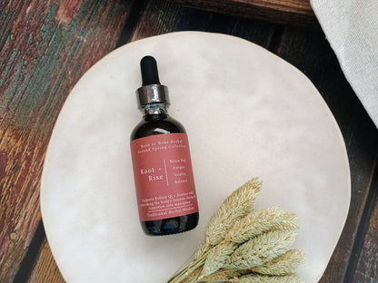 A small amber glass dropper bottle with a dusty rose label reading Root + Rise, placed on a round white plate with dried wheat stems on a rustic wooden surface.