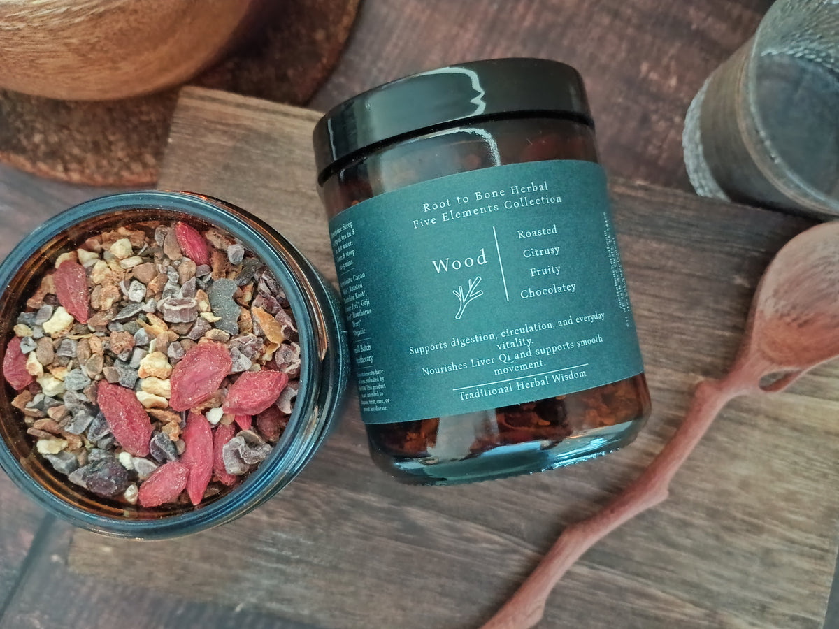 An open glass jar of herbal tea and a closed amber jar labeled 'Wood' from Root to Bone Herbal Collection, styled on a rustic wooden surface with a wooden spoon and a coconut bowl in the background.