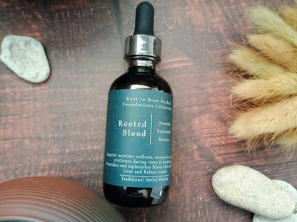 A dark amber glass dropper bottle labeled 'Rooted Blood' by Root to Root Herbal Foundations Collection, placed on a rustic wooden surface with decorative stones and dried grass.