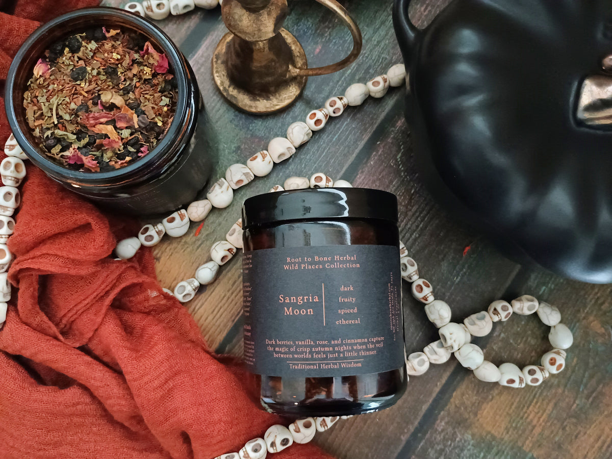 A dark amber glass jar of 'Sangria Moon' herbal blend by Root to Bone Herbal, styled on a rustic wooden background with a red cloth, skull beads, a brass candle holder, and a black teapot.