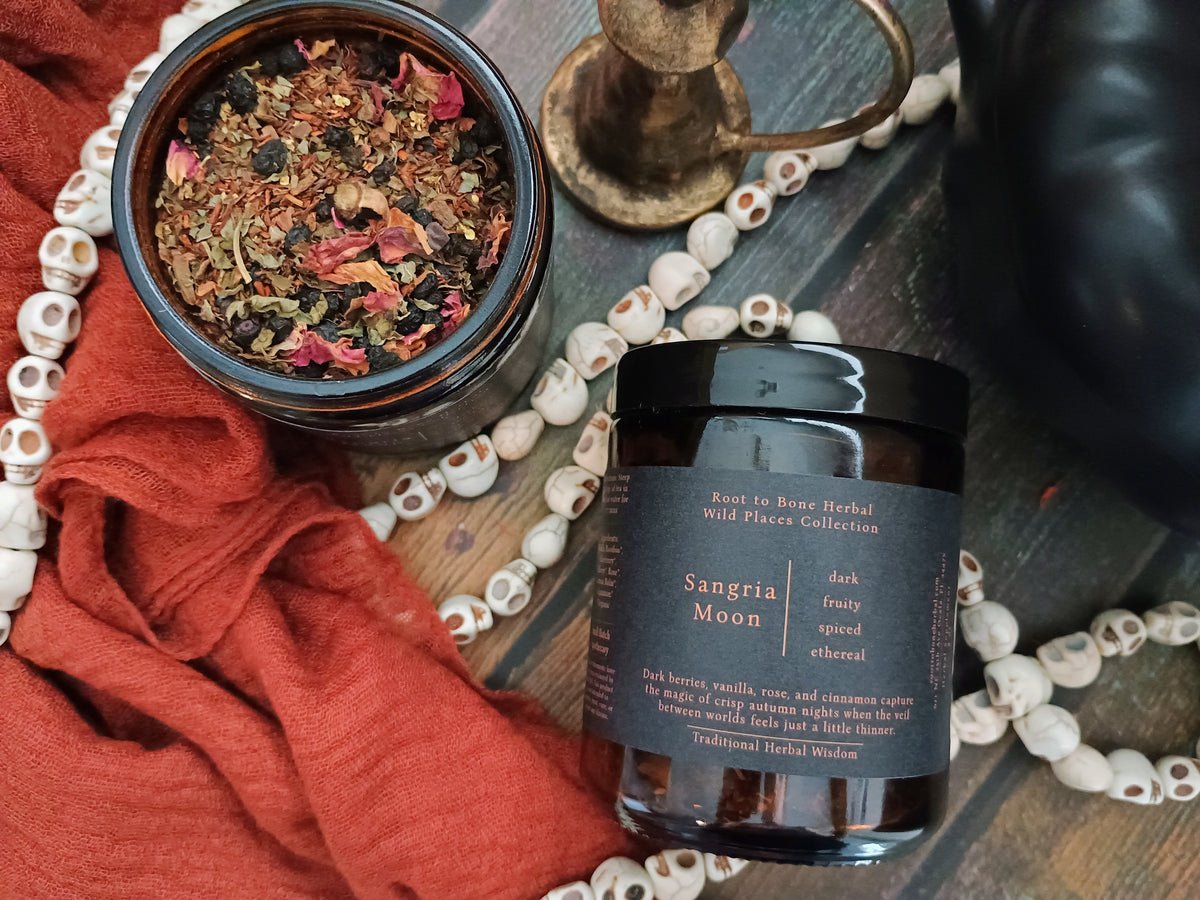 A jar of Sangria Moon herbal blend shown next to an open jar displaying the dried flower and herb blend inside. The scene is styled with a rust-colored cloth, a white skull bead necklace, and a brass candlestick on a dark wooden surface.