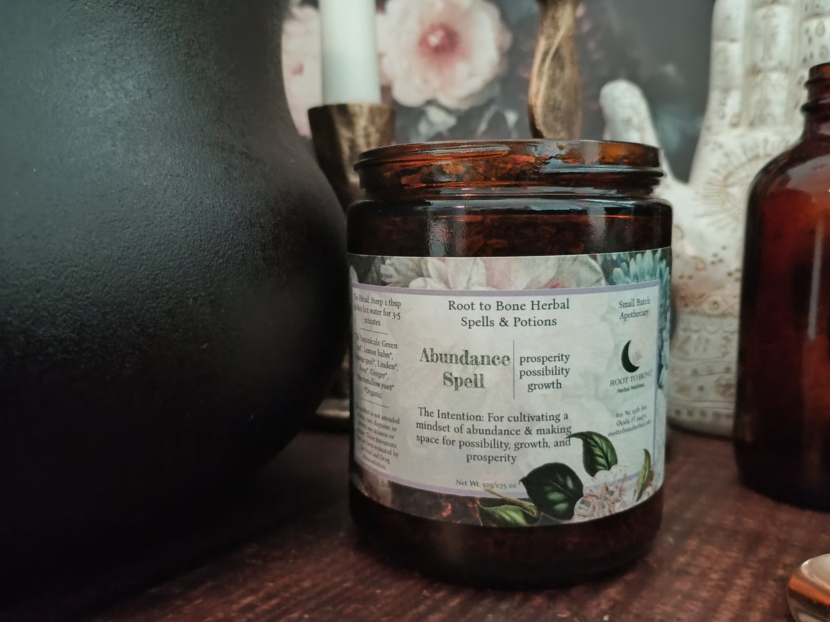 An amber glass jar candle labeled 'Abundance Spell' by Root to Bone Herbal Spells & Potions, styled on a wooden surface with mystical props including a black ceramic vessel, a white candle, and another amber bottle.