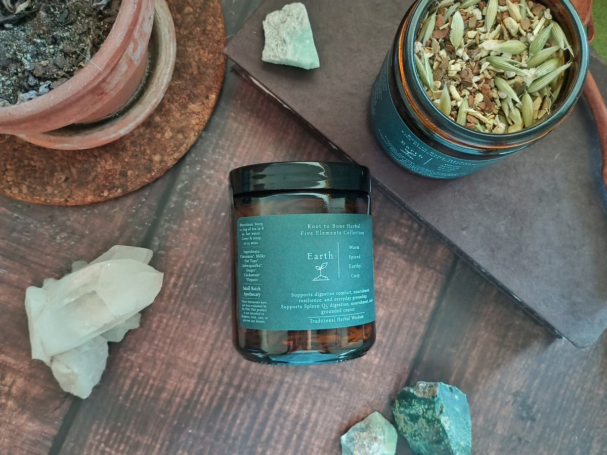A glass jar with a dark teal label and black lid sits on a wooden surface, surrounded by crystals, a potted plant, and another open jar of dried botanicals.