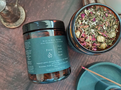 An amber glass jar of Root to Bone Herbal 'Fire' blend with a dark teal label, shown next to an open jar displaying the dried herbal mixture inside. The scene is styled on a dark wooden surface with a teal scoop and a brass spoon in the background.