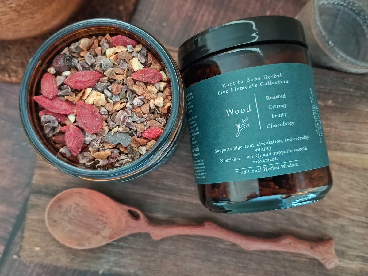 An open round glass jar filled with herbal blend and a black-lidded amber jar labeled 'Wood' from Root to Bone Herbal, placed on a rustic wooden surface with a wooden spoon nearby.