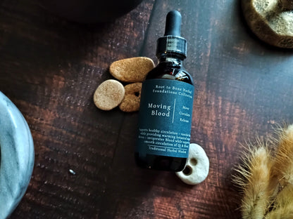 A dark amber glass dropper bottle labeled 'Moving Blood' by Root to Bone Herbs, styled on a dark wooden tabletop with natural stone and dried botanical props.