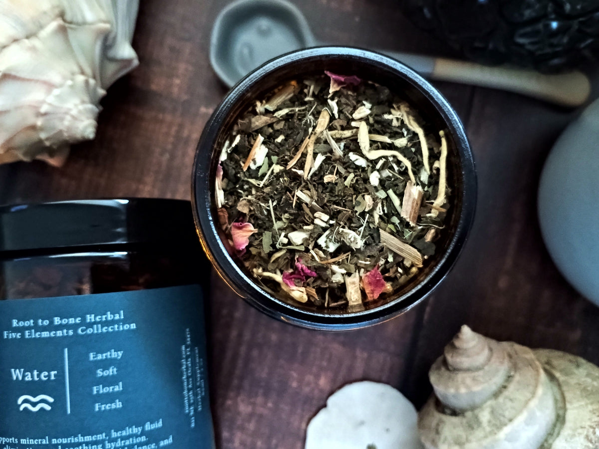An open round black jar filled with loose herbal tea blend, shown on a dark wooden surface with a branded jar and shells nearby. The label reads 'Root to Bone Herbal Five Elements Collection' and 'Water'.