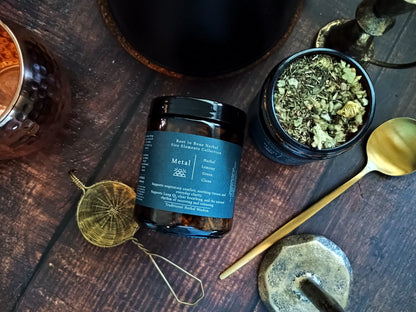 A dark amber glass jar of 'Metal' element tea by Root to Bone Herbal, styled on a dark wooden table with a gold spoon, brass tools, and an open jar showing the herbal tea inside.