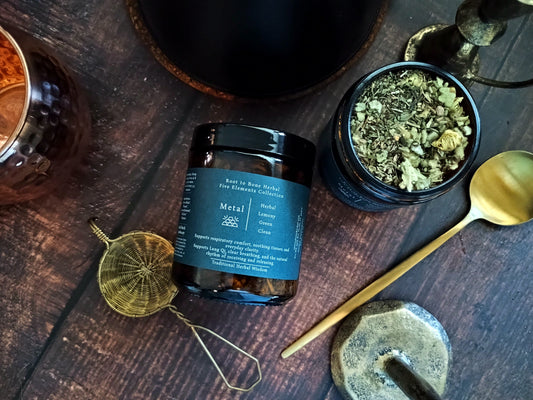 A dark amber glass jar of 'Metal' element tea by Root to Bone Herbal, styled on a dark wooden table with a gold spoon, brass tools, and an open jar showing the herbal tea inside.