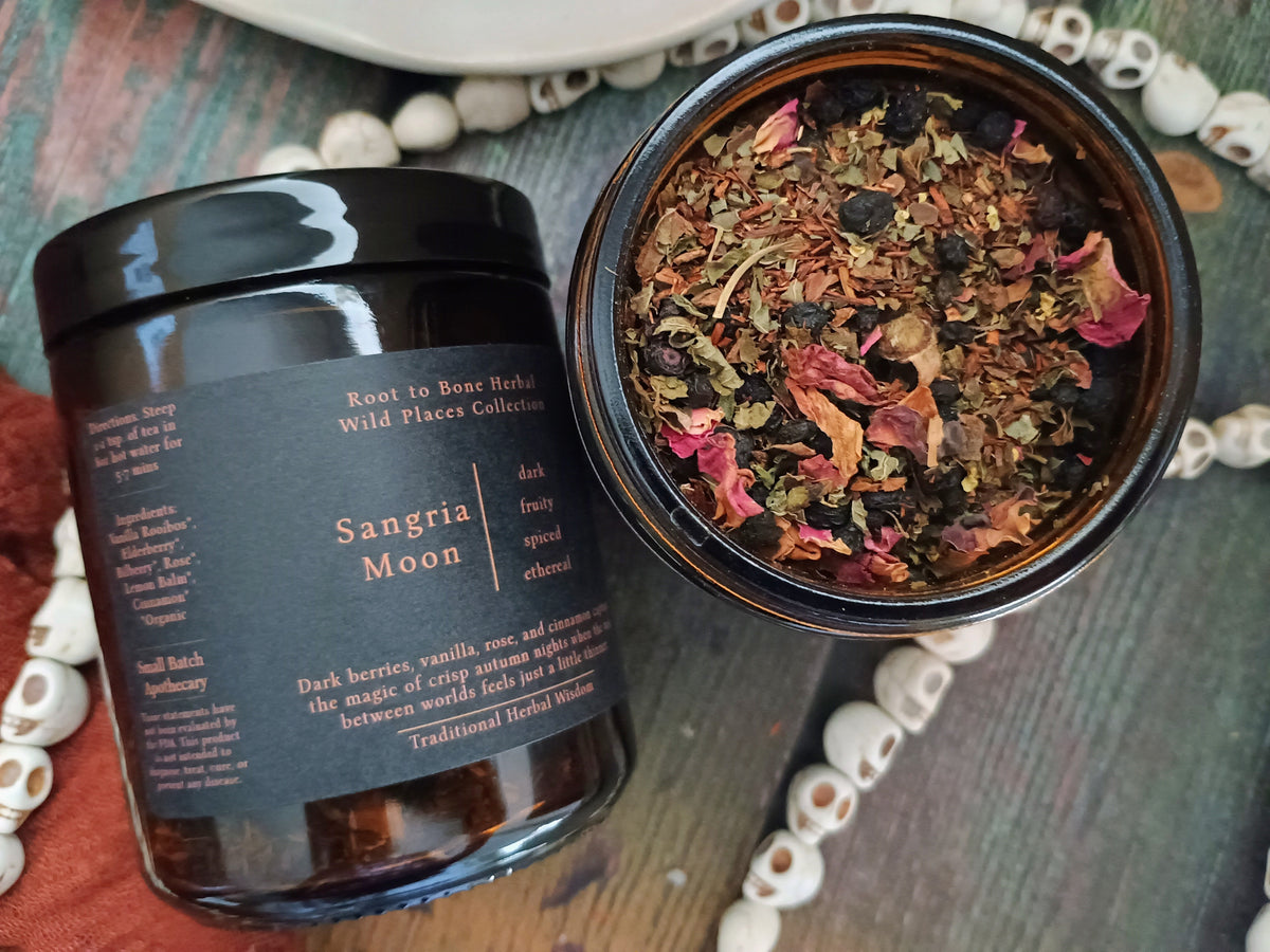 A dark glass jar labeled 'Sangria Moon' by Root to Root Herbs, shown next to an open jar displaying dried botanical ingredients like rose petals and berries, arranged on a rustic wooden surface with decorative beads.