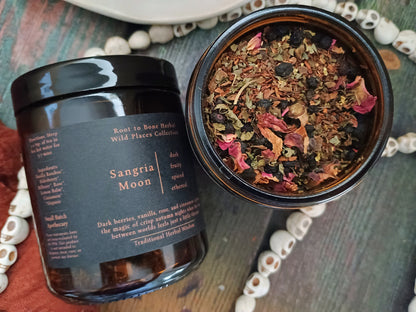 A dark glass jar labeled 'Sangria Moon' by Root to Root Herbs, shown next to an open jar displaying dried botanical ingredients like rose petals and berries, arranged on a rustic wooden surface with decorative beads.