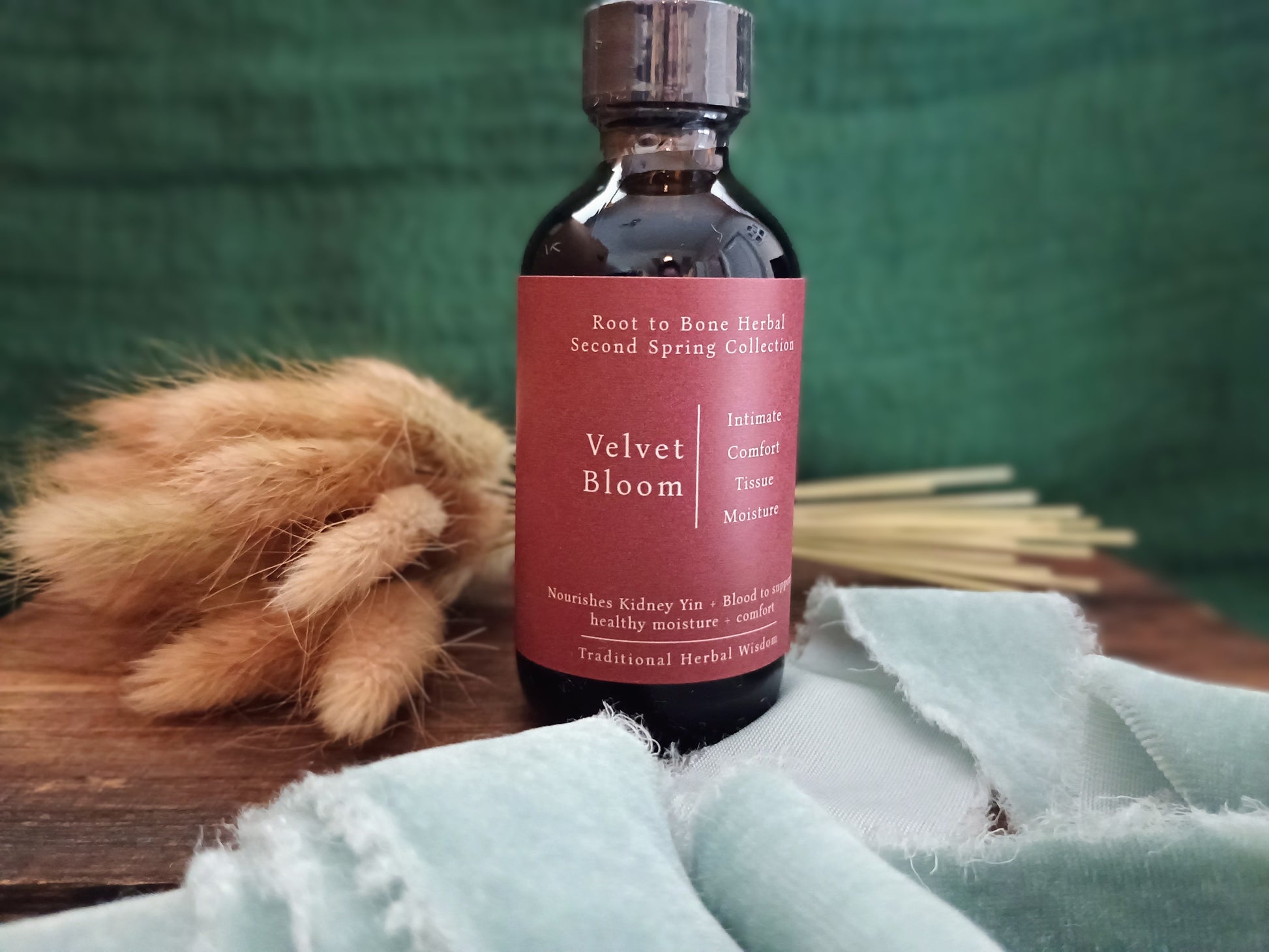 A dark amber glass bottle with a metallic cap and a dusty rose label reading 'Velvet Bloom' and 'Root to Bone Herbal Second Spring Collection,' styled on a wooden surface with a pale blue cloth and dried grasses against a dark green backdrop.