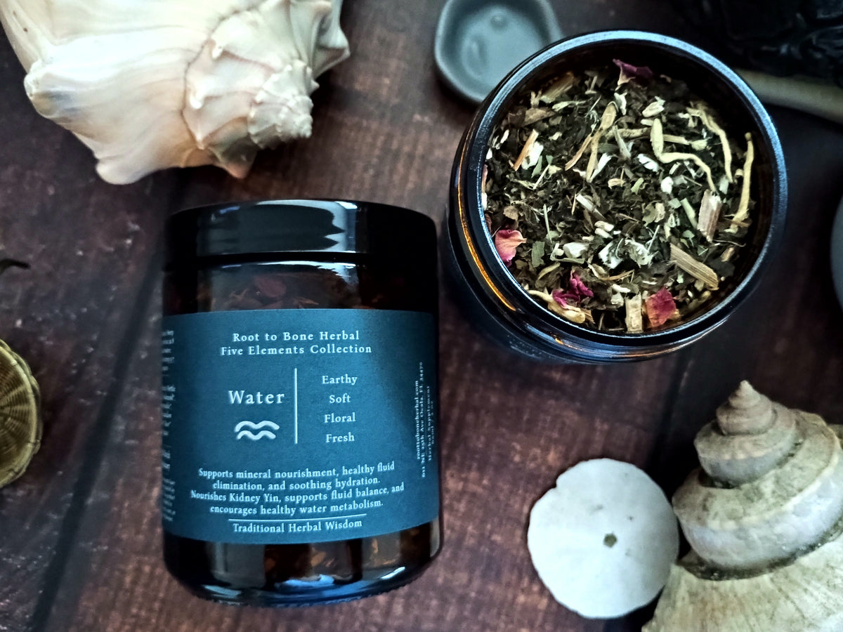 A dark amber glass jar labeled 'Water' by Root to Bone Herbal, shown next to an open jar displaying loose herbal tea blend, styled on a dark wooden surface with shells.