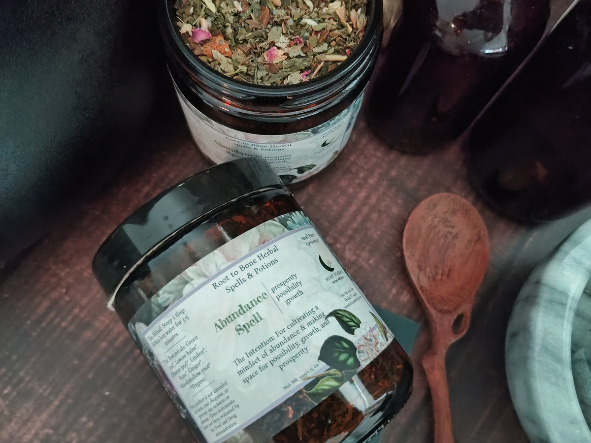 A jar of Abundance Spell herbal tea in the foreground, with another open jar of loose herbal blend behind it and a wooden spoon on a dark wooden surface.