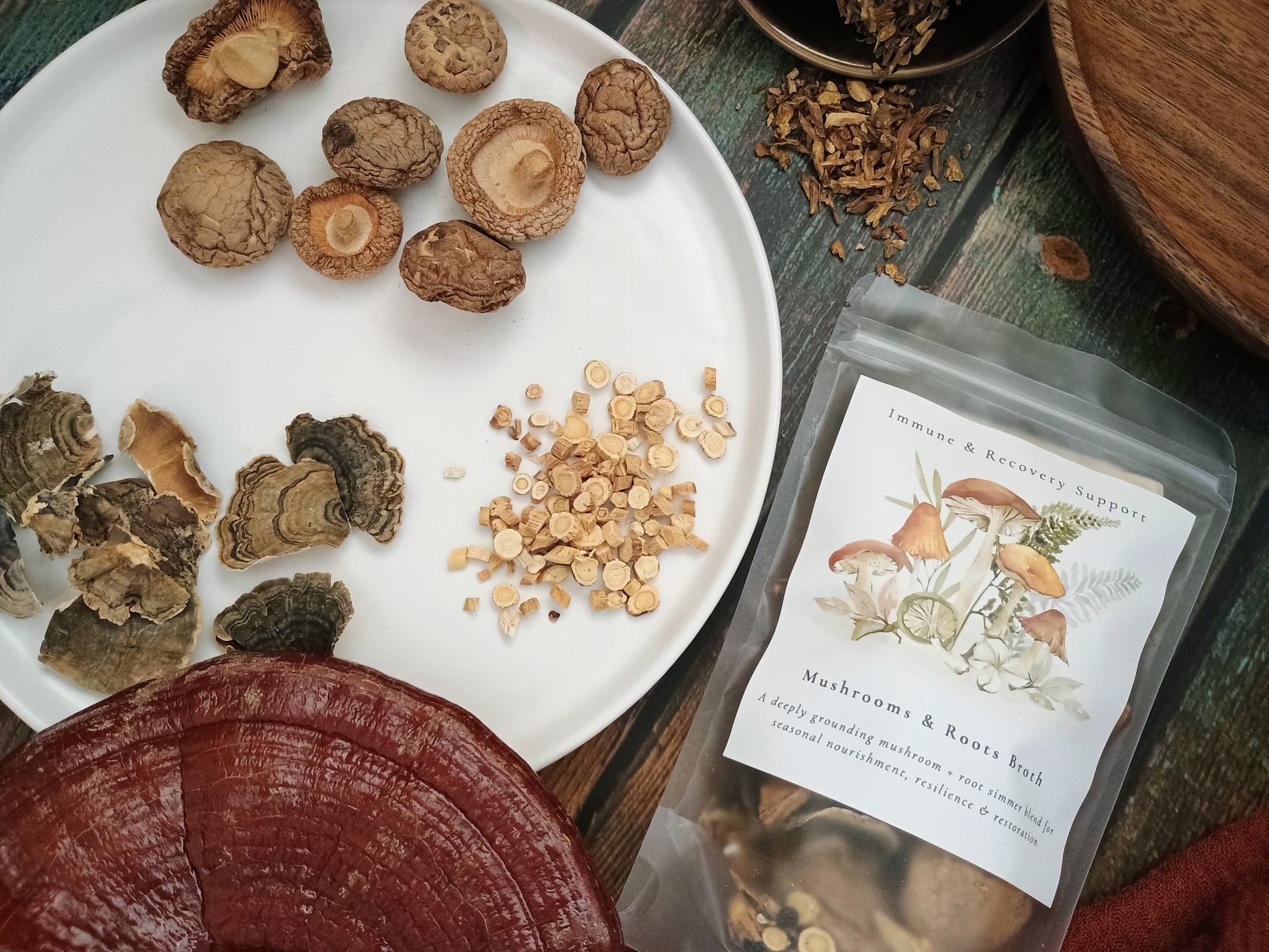 A clear stand-up pouch labeled 'Mushrooms & Root Edamame' with illustrated mushroom artwork, placed on a rustic wooden surface beside a white plate of dried mushrooms, sliced mushrooms, and small dried roots. The scene is styled as a flat lay with natural ingredients and wood props.