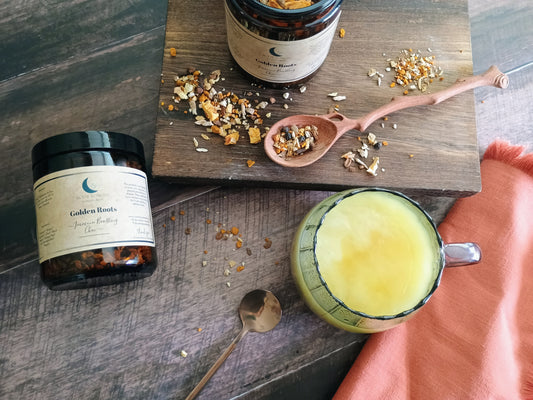 Two jars of 'Golden Roots' on a wooden surface with a glass of turmeric chai latte and a spoon.