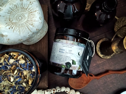 A jar of Root to Bone Herbal Spells & Potions 'Clarity Seeker' tea, styled on a dark wooden surface with dried herbs, a carved stone, and brass tools.
