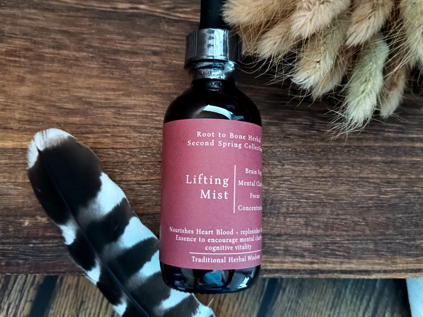 A dark amber glass bottle of Lifting Mist herbal essence with a black dropper cap and a dusty rose label, displayed on a rustic wooden surface with a black and white feather and dried grass as props.