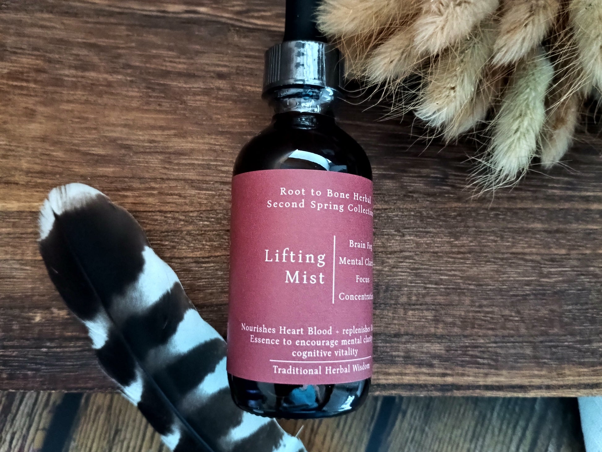 A dark amber glass bottle of Lifting Mist herbal essence with a black dropper cap and a dusty rose label, displayed on a rustic wooden surface with a black and white feather and dried grass as props.