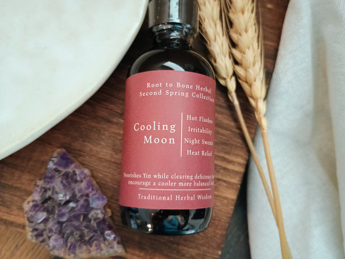 A dark glass bottle of Cooling Moon herbal tincture by Root to Bone Herbal, featuring a dusty rose pink label, styled on a wooden surface with wheat stalks, a white ceramic dish, and a purple crystal cluster.