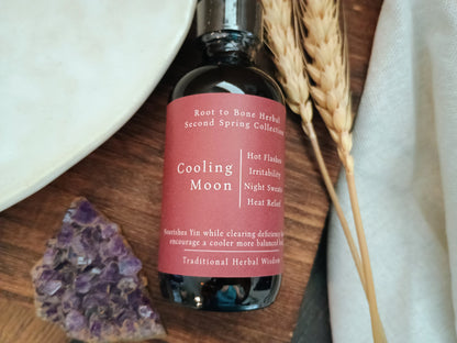 A dark glass bottle of Cooling Moon herbal tincture by Root to Bone Herbal, featuring a dusty rose pink label, styled on a wooden surface with wheat stalks, a white ceramic dish, and a purple crystal cluster.