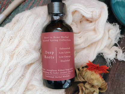 An amber glass bottle of Root to Bone Herbal Deep Roots tincture with a black dropper cap, resting on a bed of cream-colored silk or gauze with dried botanical elements.