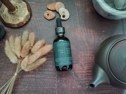 A dark amber glass dropper bottle labeled 'Deep Yin' by Root to Bone Herbs, styled on a dark wooden surface with dried bunny tail grass, small stones, a metal stand, and a dark ceramic teapot.
