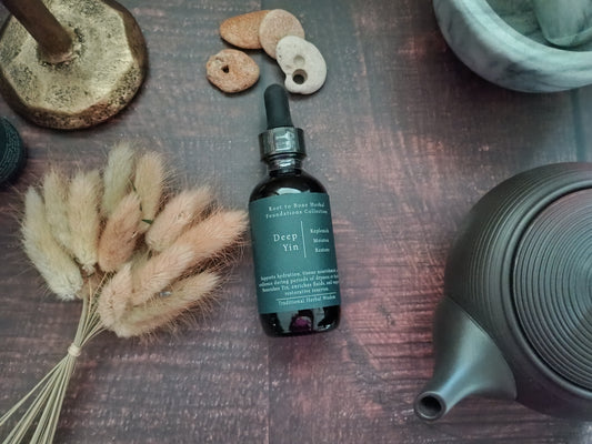 A dark amber glass dropper bottle labeled 'Deep Yin' by Root to Bone Herbs, styled on a dark wooden surface with dried bunny tail grass, small stones, a metal stand, and a dark ceramic teapot.