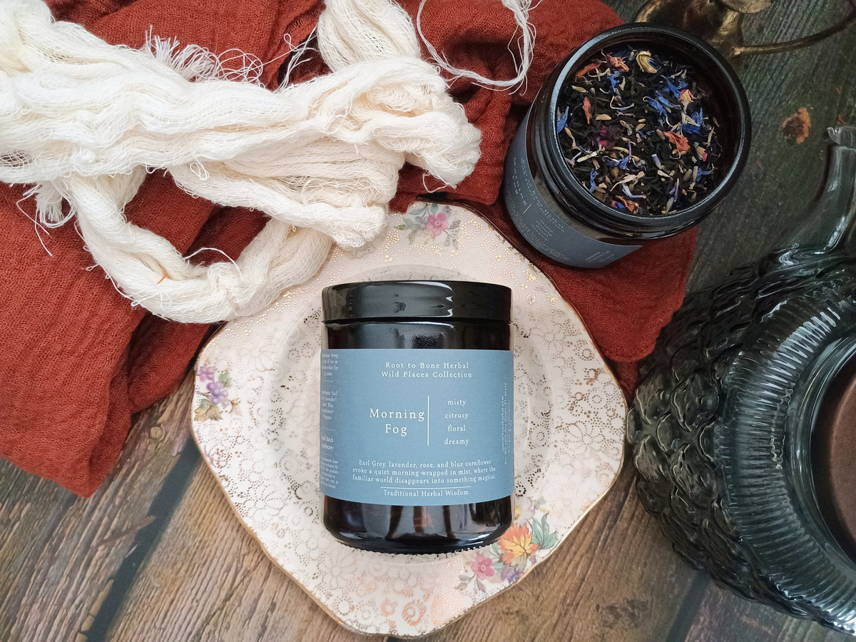 A dark amber jar labeled 'Morning Fog' by Root to Bone Herbal, styled on a vintage floral plate with a rust-colored cloth, cream yarn, and a glass teapot on a wooden table.