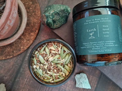 An open jar of Earth to Bone Herbal Five Elements Collection spiced earthy cozy herbal tea, showing the dried herb blend inside, next to its dark amber glass jar with a dark green label, styled on a wooden surface with crystals.