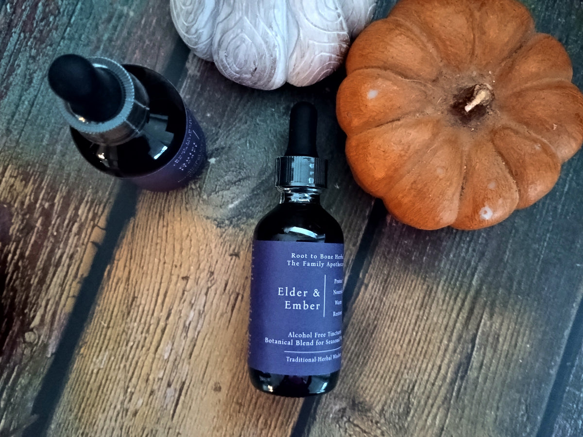 A bottle of Elder & Ember herbal tincture with a purple label and black dropper cap, styled on a rustic wooden surface next to a small orange pumpkin, a white textured ball, and another dark bottle.
