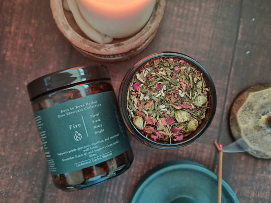 A dark amber glass jar of Root to Bone Herbs 'Fire' blend with a black lid, shown next to an open jar displaying the dried herbal mixture inside. The scene is styled on a dark wooden surface with a lit candle in a wooden bowl, a match, and a stone weight.