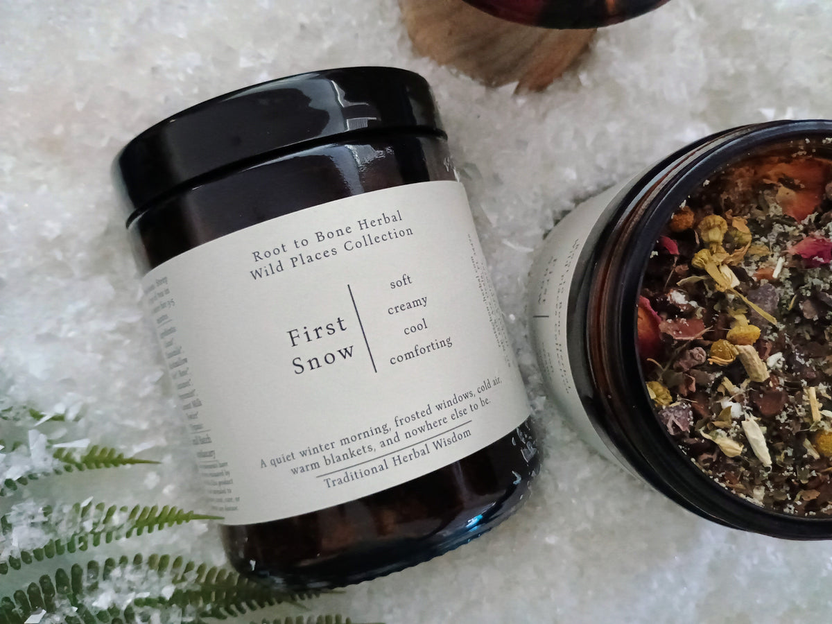 Two amber glass jars of First Snow Herbal Wild Places Collection herbal tea on a bed of white shavings, with one jar open to show the dried botanical blend inside.