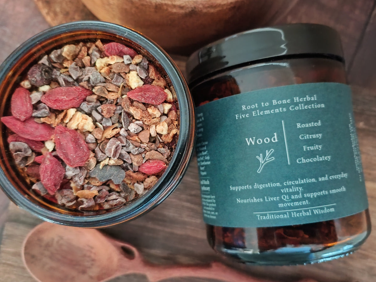 An open jar of herbal tea and a closed jar of the same product on a wooden surface. The tea contains dark chocolate pieces, brown roots, and red dried berries.
