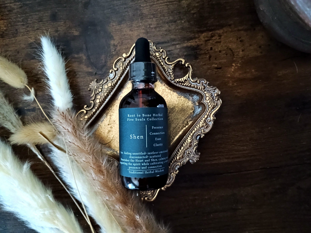 A dark amber glass dropper bottle labeled 'Root to Bone Herbs Five Souls Collection Shen' resting on an ornate gold tray over a dark wooden surface, styled with dried pampas grass.