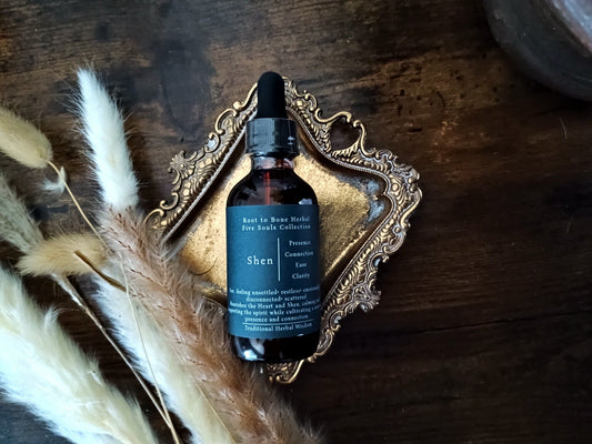 A dark amber glass dropper bottle labeled 'Root to Bone Herbs Five Souls Collection Shen' resting on an ornate gold tray over a dark wooden surface, styled with dried pampas grass.
