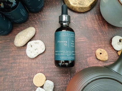 A dark amber glass dropper bottle labeled 'Flowing Qi' by Root to Root Herbs, styled on a dark wooden surface with decorative stones and a teapot.