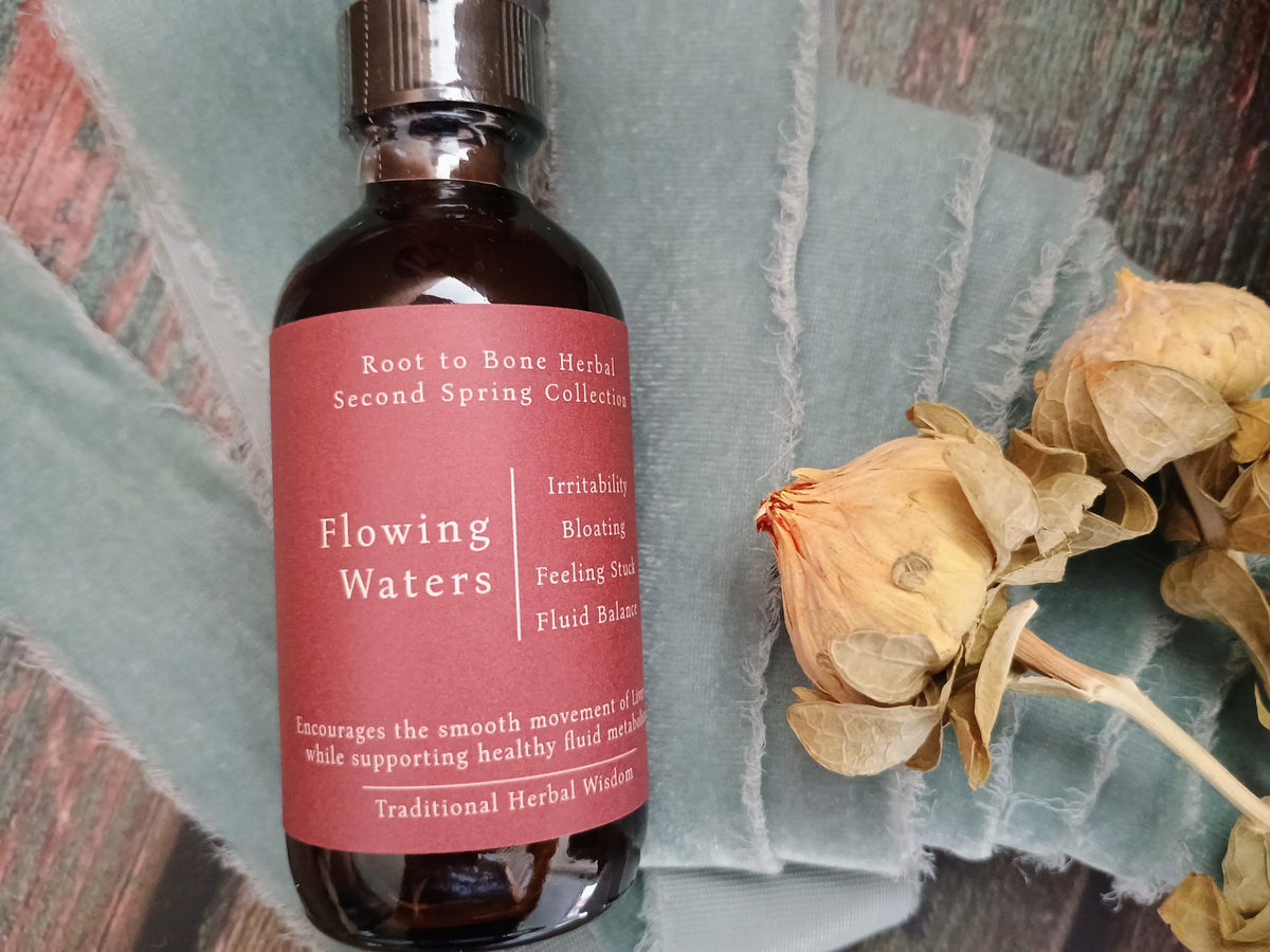A bottle of Root to Bone Herbal Second Spring Collection 'Flowing Waters' herbal liquid, featuring a dusty rose label and dark amber glass, styled on a textured teal fabric next to dried roses.