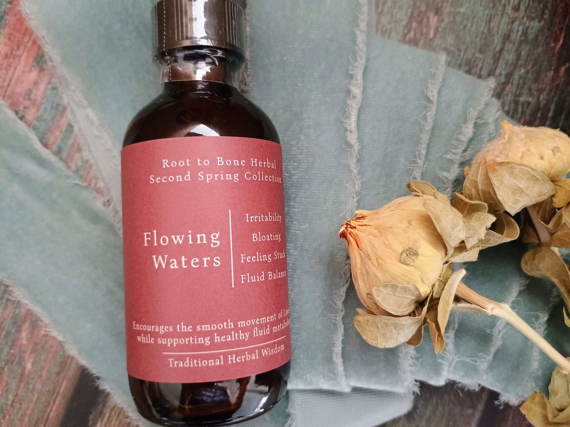 A bottle of Root to Bone Herbal Second Spring Collection 'Flowing Waters' herbal liquid, featuring a dusty rose label and dark amber glass, styled on a textured teal fabric next to dried roses.