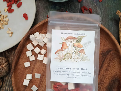 A clear stand-up pouch labeled 'Restorative Nourishment' and 'Nourishing Earth Blend' sits on a wooden tray, surrounded by small white herbal cubes, red goji berries, and dried botanicals on a rustic dark wood surface.
