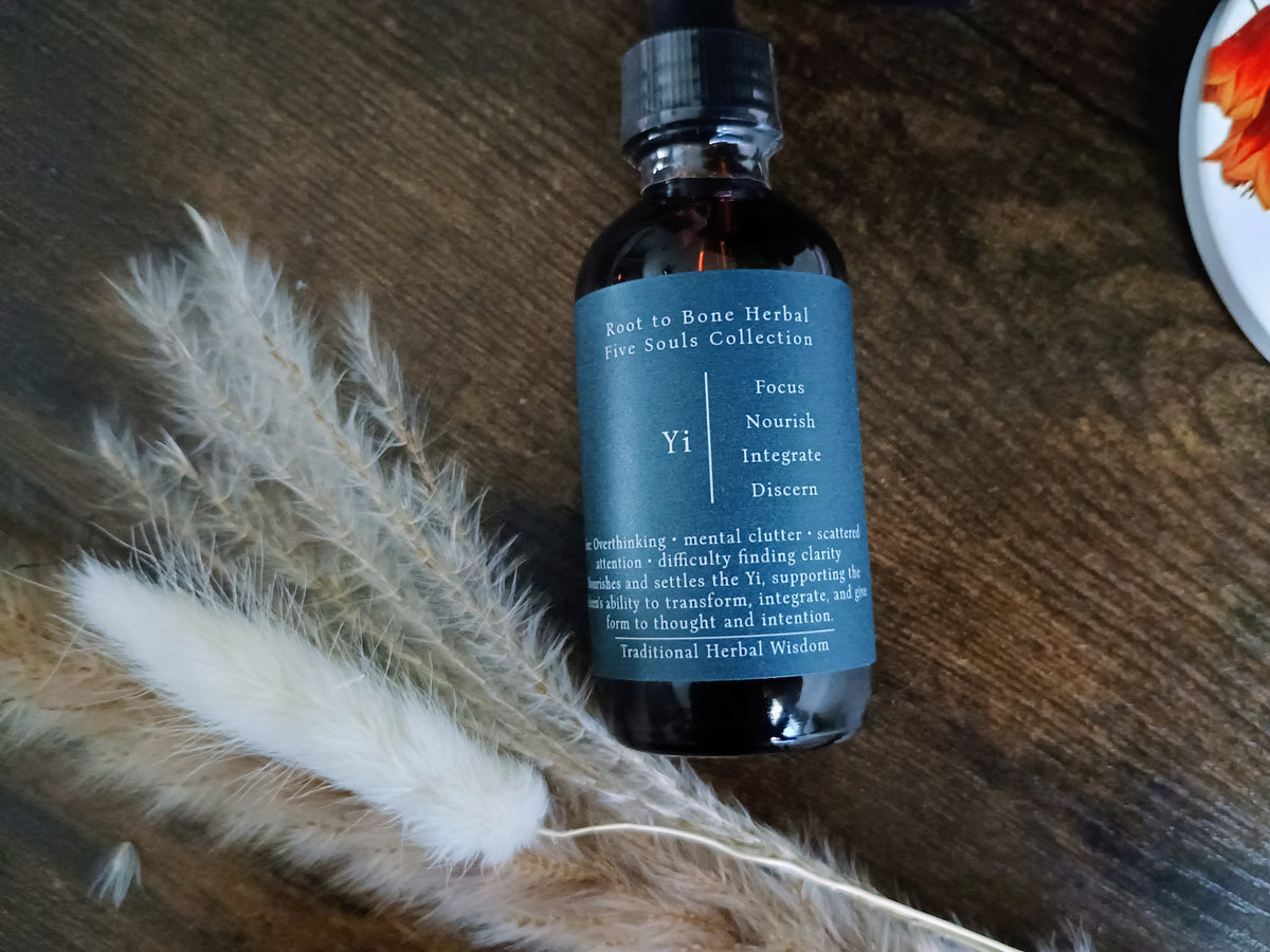 A dark amber glass bottle with a black cap and a dark slate-blue label reading 'Root to Bone Herbal Five Senses Collection Yi', styled on a dark wooden surface with dried pampas grass and a partial white plate in the corner.