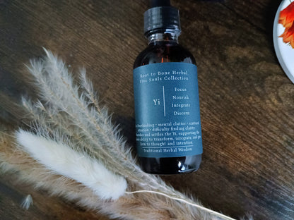 A dark amber glass bottle with a black cap and a dark slate-blue label reading 'Root to Bone Herbal Five Senses Collection Yi', styled on a dark wooden surface with dried pampas grass and a partial white plate in the corner.