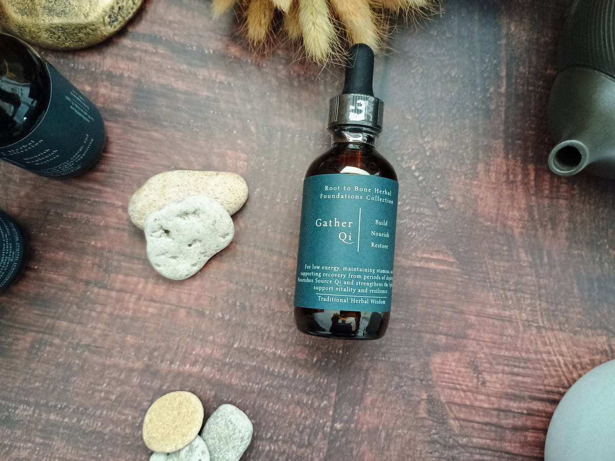 A dark amber glass dropper bottle labeled 'Gather Qi' by Root to Bone Apothecary, styled on a dark wooden surface with natural stones and dried botanicals.