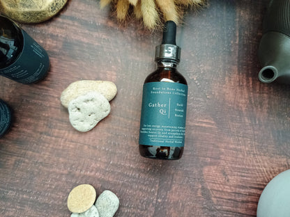 A dark amber glass dropper bottle labeled 'Gather Qi' by Root to Bone Apothecary, styled on a dark wooden surface with natural stones and dried botanicals.