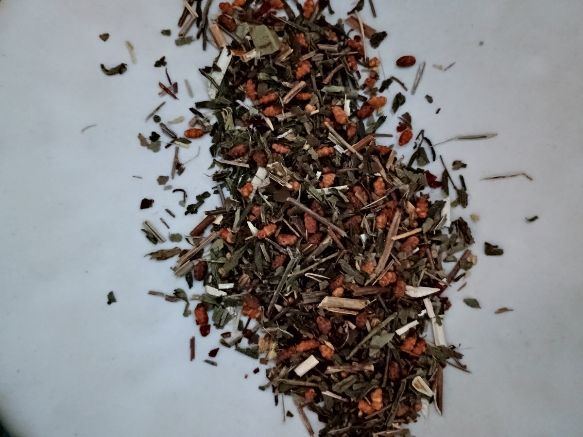 A small pile of loose dried herbal tea blend containing green, brown, and reddish-orange botanical pieces on a plain white surface.