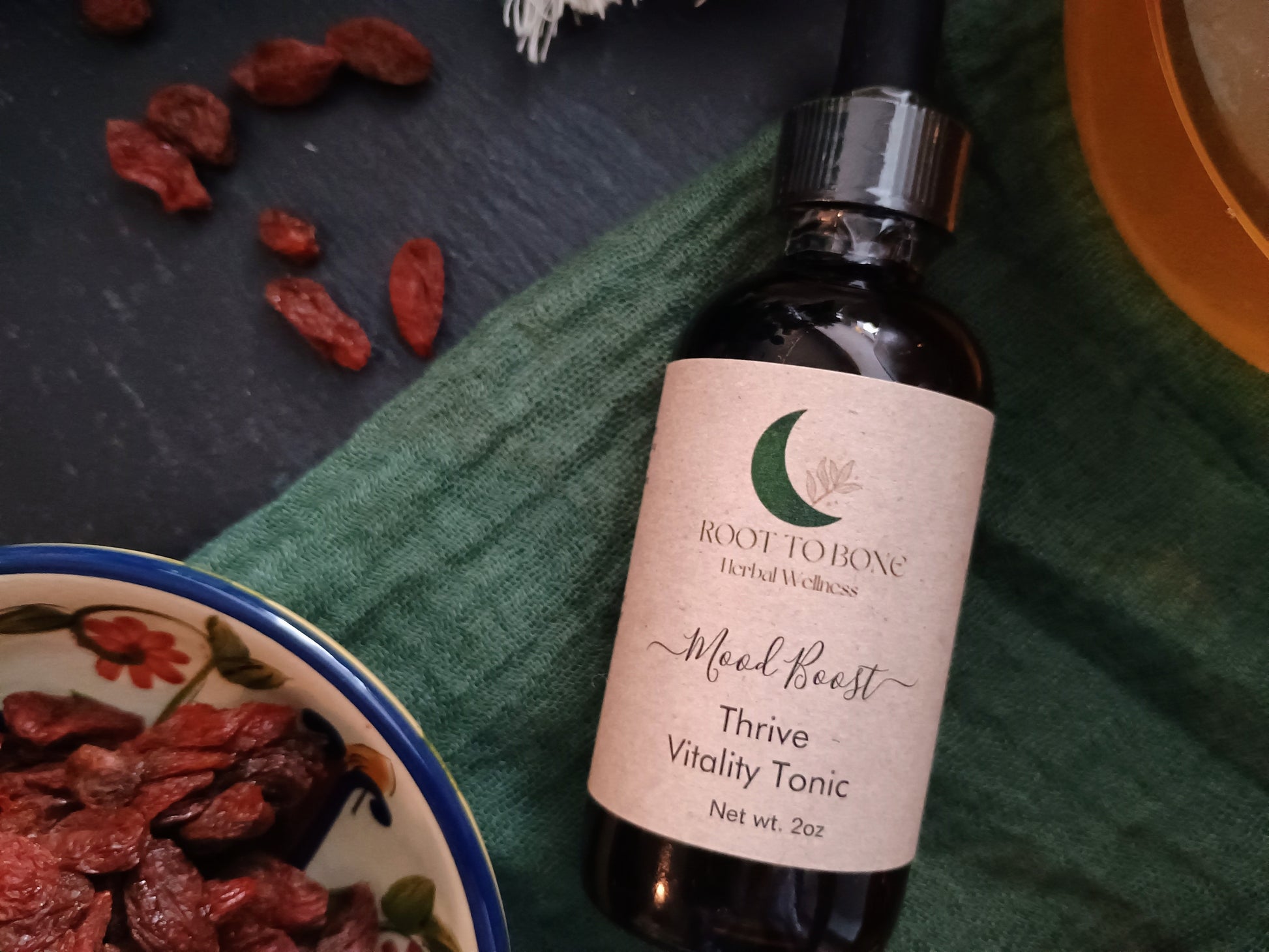 Bottle of 'Root to Bone Herbal Wellness' tonic with dried herbs on a green cloth.