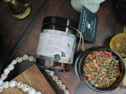 A glass jar of Root to Bone Herbal  Wellness blend with a black lid and white label, shown next to an open jar displaying the colorful herbal mixture inside. The scene is styled on a dark wooden surface with a beaded necklace, a brass object, and a small black hanging card.