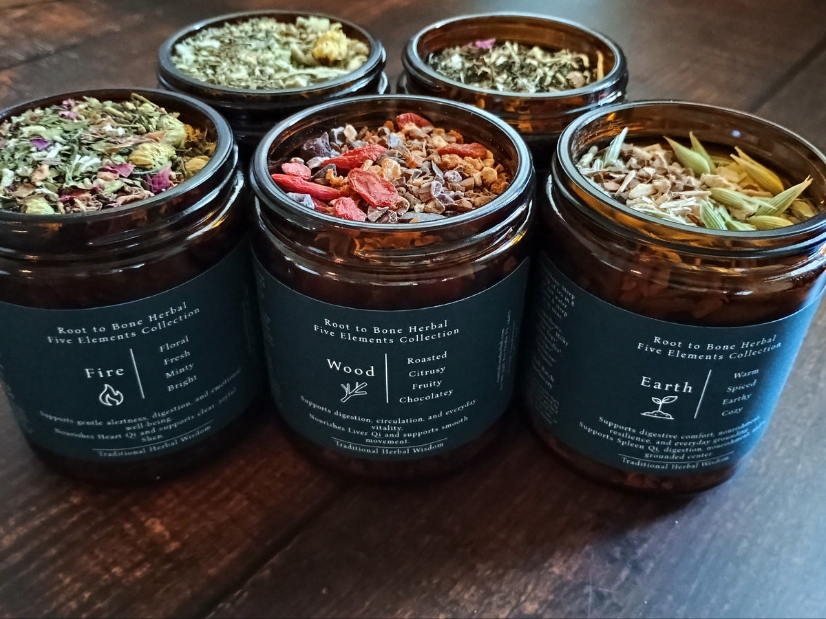 Four amber glass jars of Root to Bone Herbal Five Elements Collection teas arranged on a dark wooden table, showing their dried botanical blends on top.
