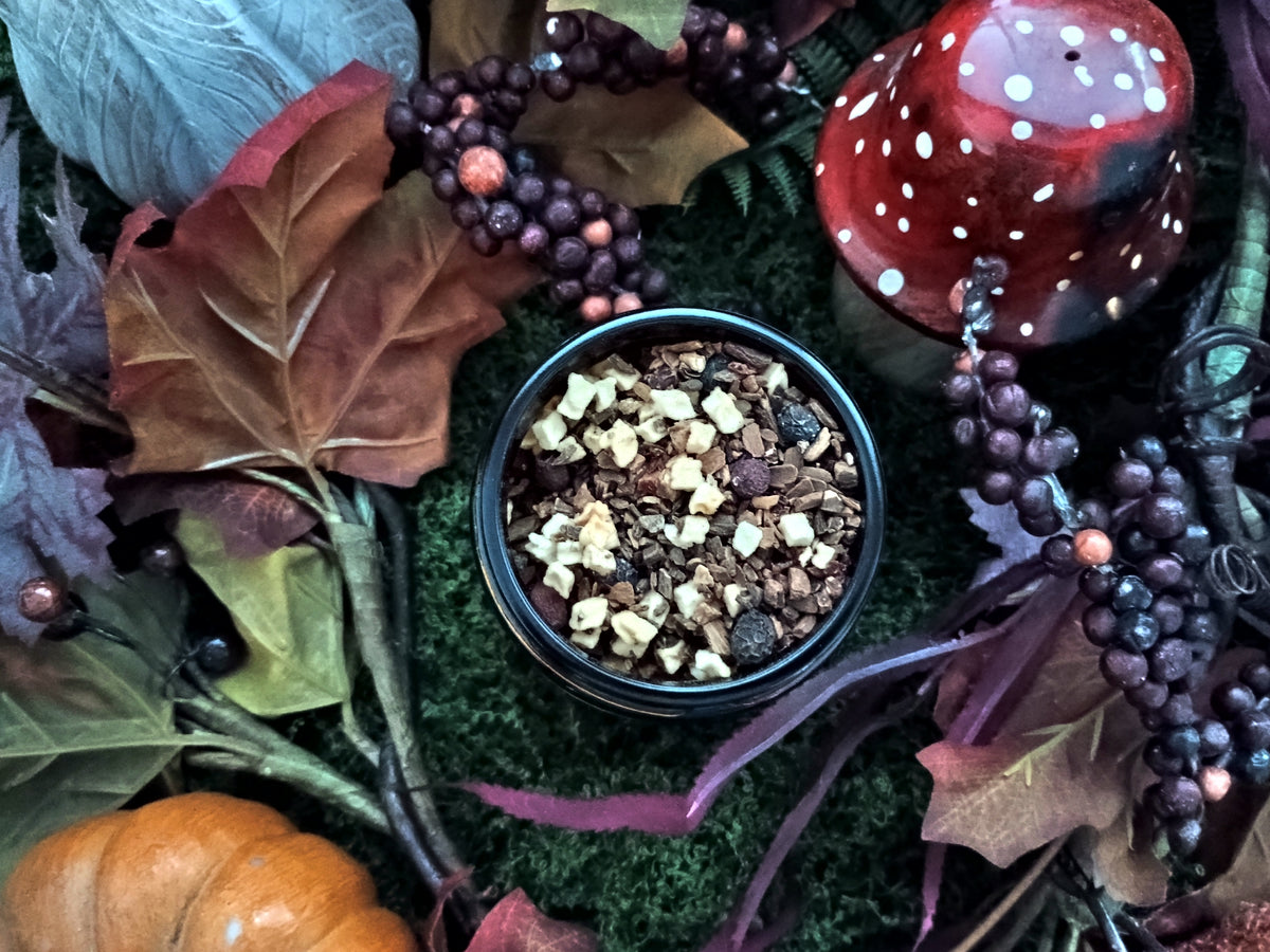 A small round black tin filled with loose leaf herbal teat, styled in a fall-themed arrangement with autumn leaves, berries, moss, a mushroom decoration, and a small orange pumpkin.