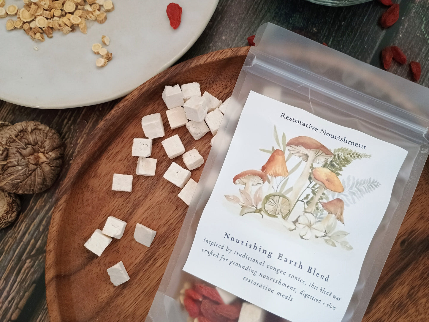 A clear stand-up pouch labeled 'Restorative Nourishment Nourishing Earth Blend' sits on a wooden board with small white cube-shaped pieces scattered around it. The scene includes dried herbs, a mushroom, and a plate in a styled tabletop setup.